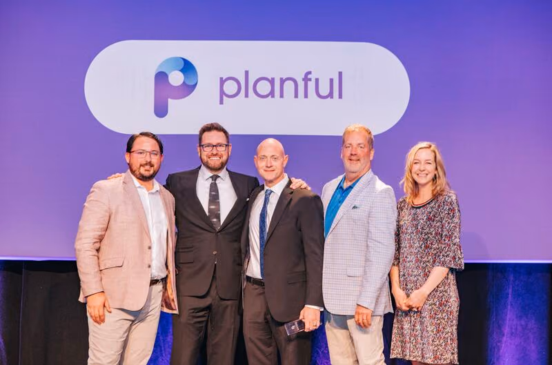 Five professionally dressed people standing together and smiling in front of a purple backdrop displaying the Planful logo.