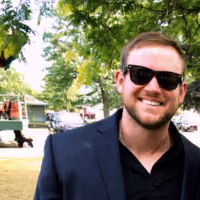 Smiling man wearing sunglasses and a dark blazer outdoors with trees and parked cars in the background.