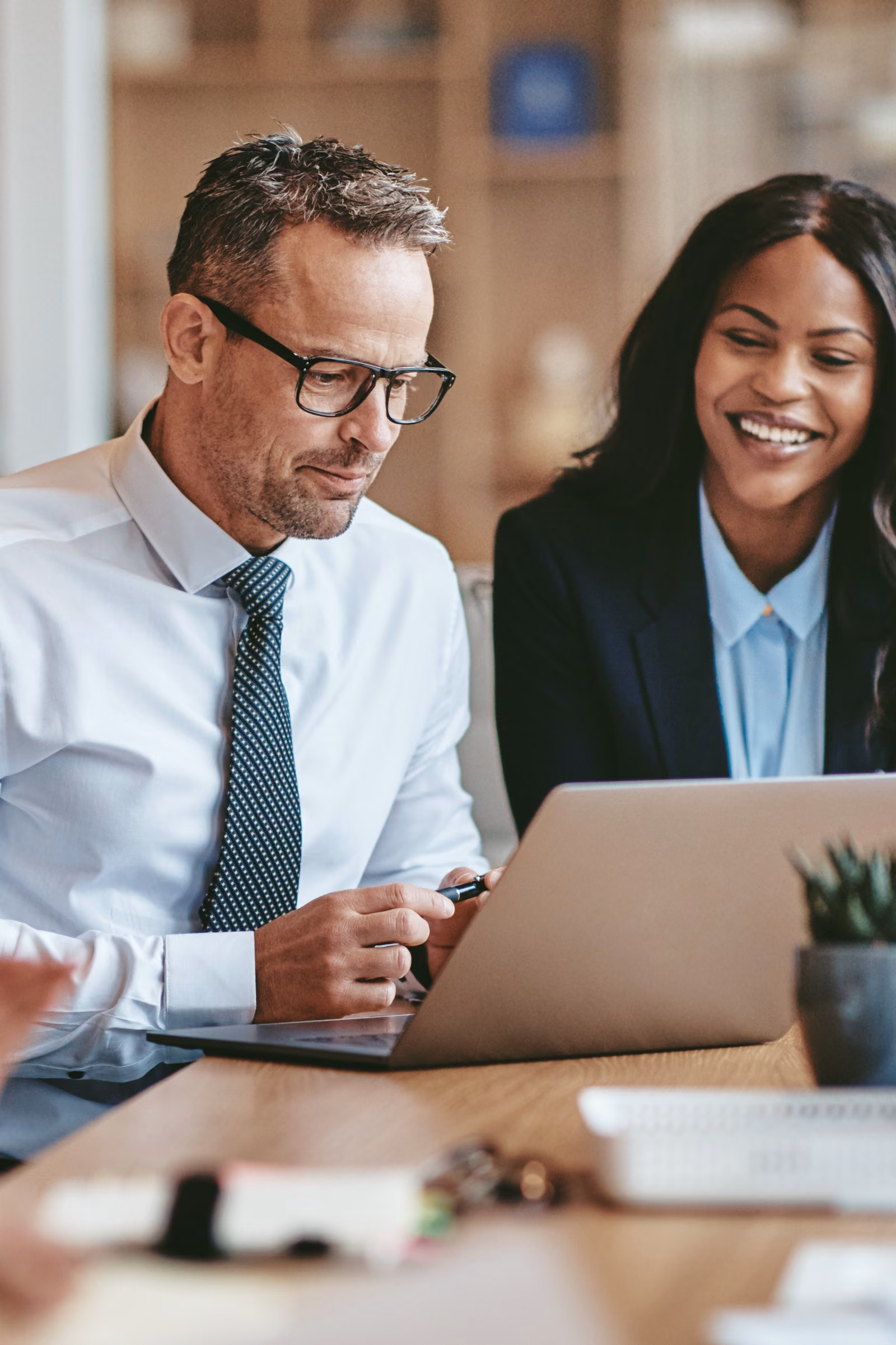 Two business professionals, a man and a woman, looking and smiling at a laptop screen together in an office setting.