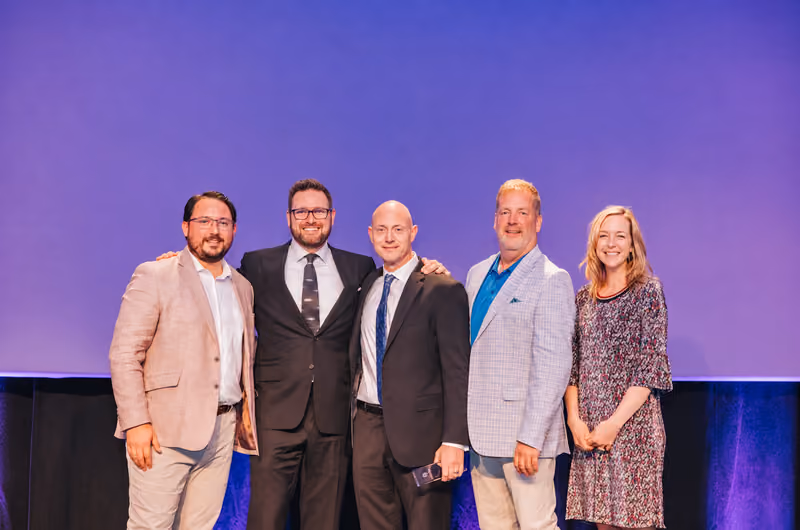 Five colleagues in business attire standing together and smiling in front of a purple stage backdrop.